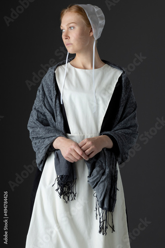 An Amish woman wearing a black dress, white apron, grey shawl, and cap against a grey backdrop