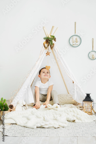 Little Caucasian boy is sitting in a toy white wigwam and smiling while looking at a wooden heart. Scandinavian Christmas interior of a children's room.