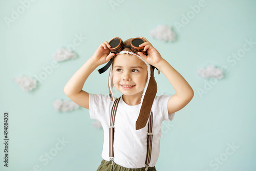 A boy in an aviator helmet and suspenders stands on a blue background with clouds and smiles.