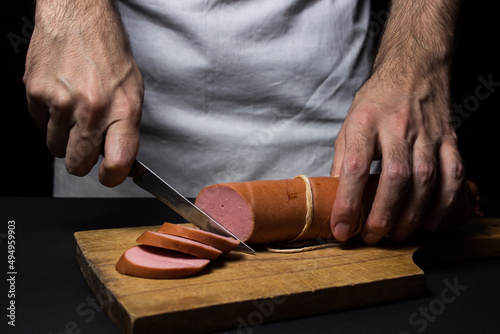 The cook cuts the sausage. Boiled sausage on a black background. Sliced sausage stick on a wooden board.