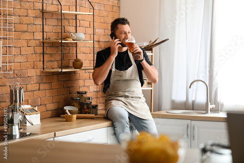 Handsome man drinking beer while talking by phone in kitchen