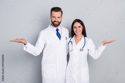 Photo of pretty happy two doctors wear white coats comparing arms empty space hugging isolated grey color background