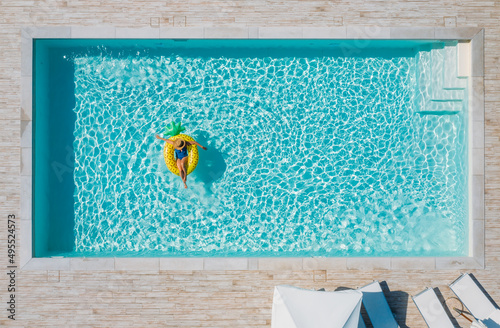 Sexy young female in swimsuit bikini, in a straw hat and sunglasses floating on blue swimming pool waves on giant inflatable Yellow Pineapple tube. Chill out summer vacation in luxury resorts concept.