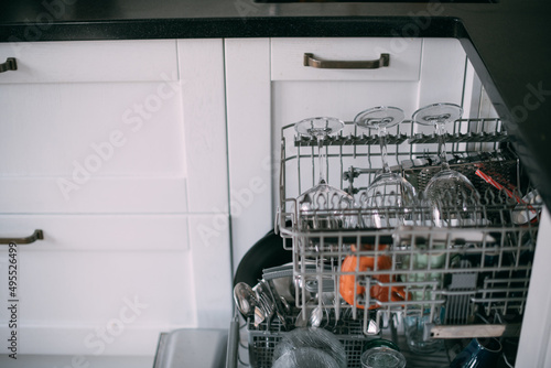 Open dishwasher with clean dishes in a white kitchen. Clean glasses, plates, mugs in the dishwasher basket