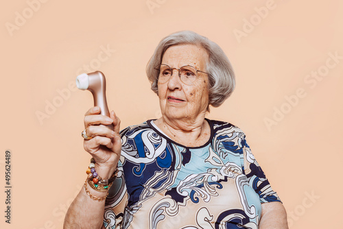 Funny doubtful elderly grandmother holding satisfyer dildo with hand at studio over beige background. Senior old woman dressing elegant looking at vibrator with funny face expression.