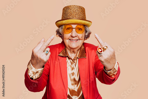 Funny stylish elderly grandmother portrait showing middle finger gesture with hand at studio. Senior old woman dressing elegant for a special event. Rebel granny over beige background.