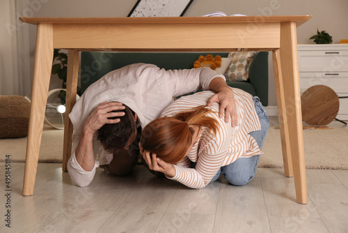 Scared couple hiding under table in living room during earthquake