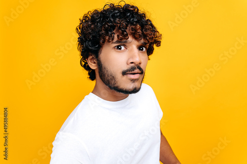 Close up of confused puzzled curly haired indian or arabian guy in white t-shirt, looking questioningly at the camera, while standing over isolated orange background