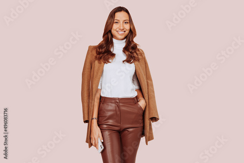 Portrait of young happy smiling businesswoman wears white turtle nec sweater, beige coat and brown leather pants, holding smart phone and posing over beige background. Business concept, copy space.