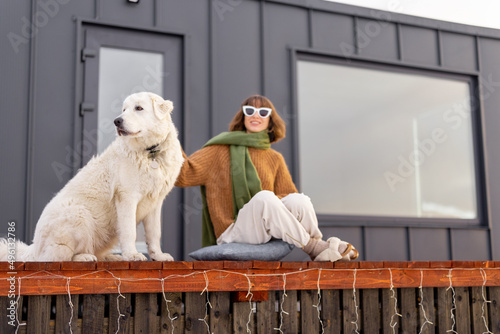 Woman sitting with dog on terrace of tiny house in the mountains. Concept of small modern cabins for rest and escape to nature. Idea of traveling with dog
