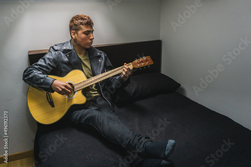 hombre tocando la guitarra sentado sobre la cama, chico joven tocando un instrumento musical en su dormitorio, chaval practicando música en su habitación 