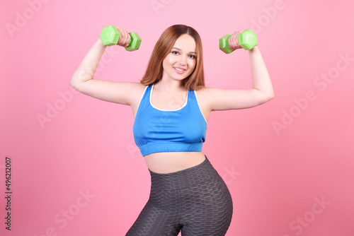 A beautiful body-positive girl on a pink background in a tracksuit lifts weights. Sports, hand training