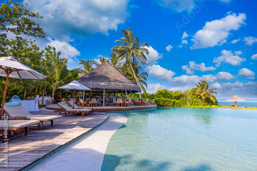 Recreational tourism landscape. Luxurious beach resort with spa swimming pool and beach chairs or leisure loungers under umbrellas with palm trees sky. Summer travel vacation background. Wellbeing