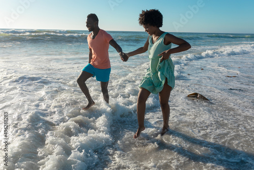 Happy african american couple enjoying summer holiday wading on shore at beach during sunny day