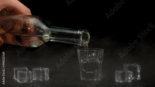 Bartender pouring up shot of vodka with ice cubes from bottle into drinking glass against black background. Barman pour clear transparent alcohol drink vodka tequila in shot-glass
