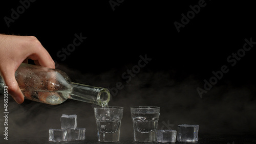 Bartender pouring up two shots of vodka with ice cubes from a bottle into glasses against black background. Barman pour clear transparent alcohol drink vodka tequila in shot-glass