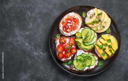 Assorted sandwiches with soft cheese, vegetables and fruits on a dark background. Top view, copy space.