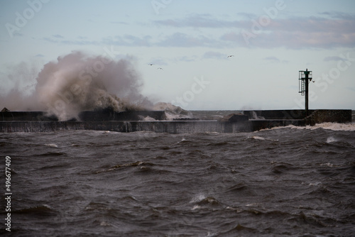 Olbrzymi sztormowy bałwan morski rozbija się o falochron. Widać ślady zniszczenia części falochronu przez żywioł morski.