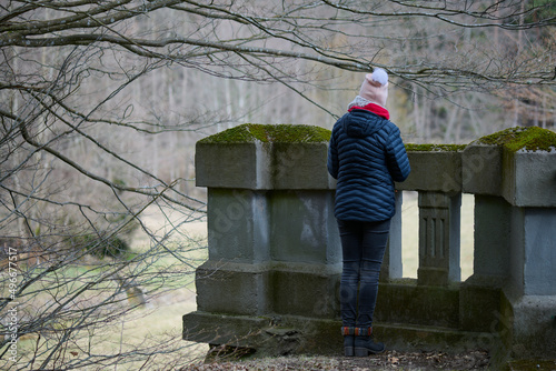 Dziewczyna odwrócona tyłem stoi przy murowanej platformie widokowej pokrytej mchem. Patrzy w dal na bezlistne drzewa,w poszukiwaniu oznak wiosny.