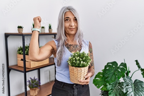 Middle age grey-haired woman holding green plant pot at home strong person showing arm muscle, confident and proud of power
