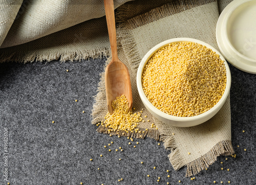 Raw organic millet grits in a white saucer with a wooden spoon on a rough napkin and a gray background.