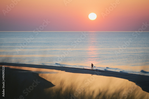 Jogging o zachodzie słońca na morskiej plaży. W tle sylwetka biegacza podczas wieczornego treningu. Kolory zachodzącego słońca, błękit, różowy, pomarańcz, czerwony, fioletowy. Odbicie słońca w wodzie.