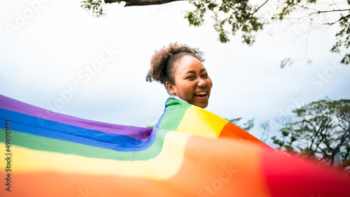 Happy young African woman carrying LGBT rainbow flag symbol and smiling in camera in green city park. Concept of homophobia,diversity,equity, peace and love,freedom,liberty,Lesbian,Feminist,Banner.