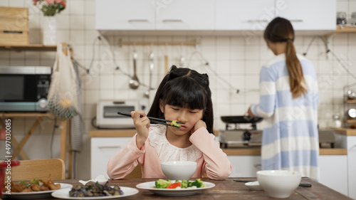 selective focus of unhappy picky Asian schoolgirl daughter holding face with disgust look on face while playing with food in bowl at dining table. her mother is cooking in kitchen at background