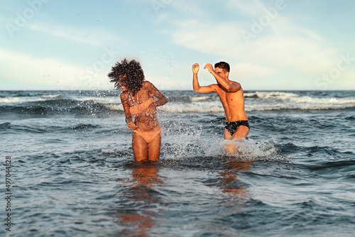 Engaged biracial couple on vacation playing together splashing water bathing in between the waves - multi ethnic young people enjoying summertime carefree
