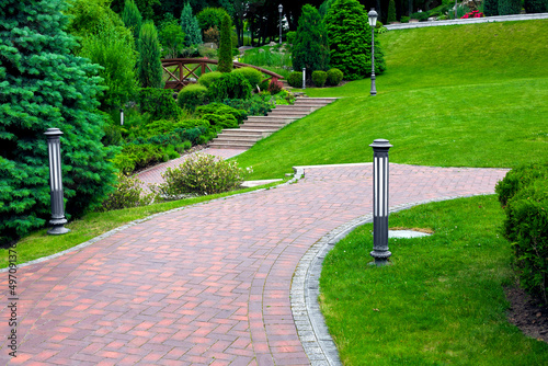 iron ground lantern and poli garden lighting of park curved path paved with stone tiles with drain lattice in park among plants, evergreen bushes and pine trees on slope hill green lawn, nobody.