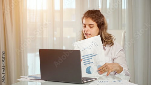 Disabled woman freelancer with infantile cerebral paralysis talks on videocall via laptop with manager showing project paper against window at home.
