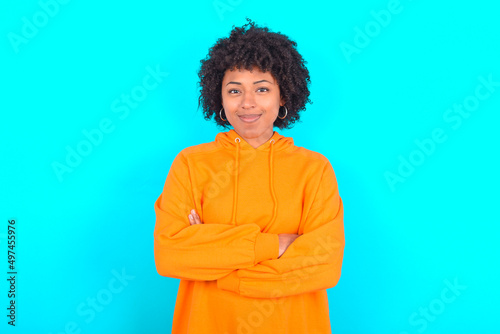 Self confident serious calm young woman with afro hairstyle wearing orange hoodie against blue background stands with arms folded. Shows professional vibe stands in assertive pose.
