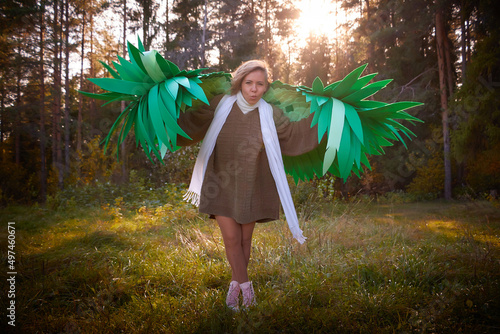 Cute girl in in a grey sweater, white scarf and green wings in nature landscape on sunny autumn. Model poses in green and yellow landscape as elf, dryad or angel. Unusual photo shoot on a fine day