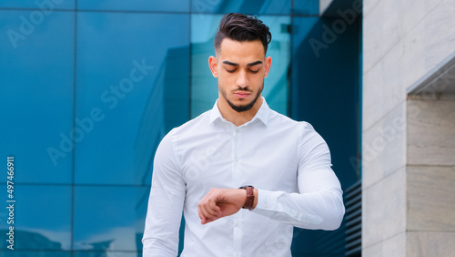 Serious upset angry hispanic indian man bearded guy disgruntled businessman boss wears formal stylish shirt stands outdoors looking at wrist watch worried about being late lateness time rush waiting