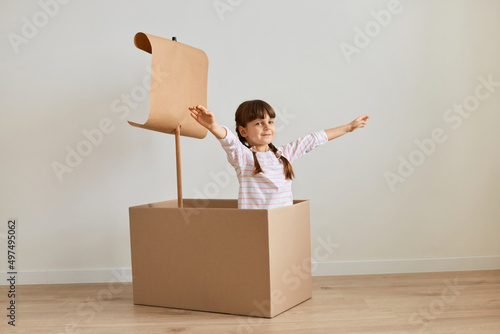 Portrait of happy positive dark haired little girl playing in handmade ship from cardboard box with sail, posing with raised arms, imagines she is sailing.