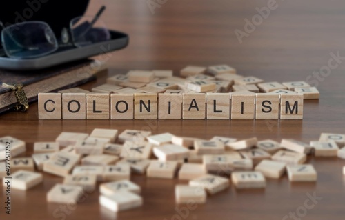 colonialism word or concept represented by wooden letter tiles on a wooden table with glasses and a book