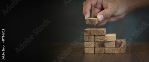 Hand is placing wood block tower stack in pyramid stair step with caution to prevent collapse or crash concepts of financial risk management and strategic planning and business challenge plan.