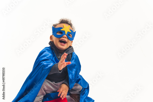 Studio shot of a little biracial boy toddler in a hero costume with a dark blue mask with a star and a cape laughing out loud over white background. High quality photo