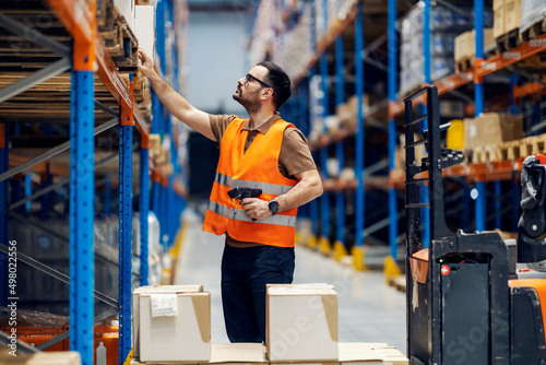 A dispatcher with scanner in hands looking at boxes on shelves in warehouse.