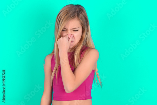 blonde little kid girl wearing pink sport clothes over green background , holding his nose because of a bad smell.