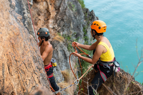 Asian man and woman climber climbing to rock mountain peak together at tropical island in sunny day. Strong male and female enjoy outdoor active lifestyle and extreme sport climbing on summer vacation