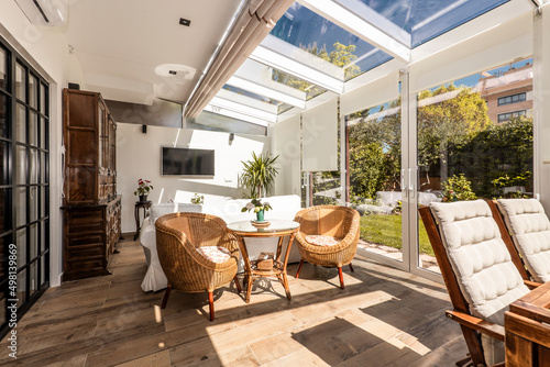 Interior of a closed white aluminum and glass terrace with large closed sliding doors with access to a garden with lawn and ornamental plants, wooden and wicker furniture inside, stoneware floors.