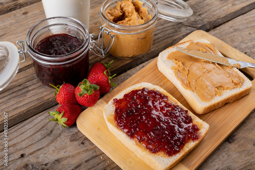 Close-up peanut butter and jelly sandwich on serving board with jars, strawberries and milk at table