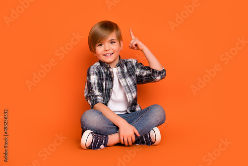 Full size photo of young boy sit floor indicate finger creative choice smart isolated over orange color background