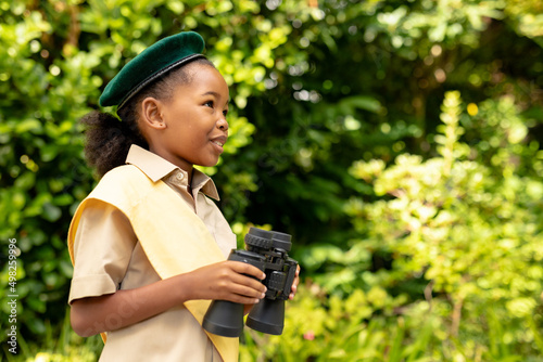 Smiling african american scout girl in uniform holding binoculars in forest