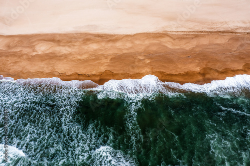 drone view of whitecapped waves crashing on an empty golden sand beach