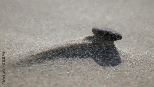 Kamień po silnej wichurze leży na utworzonej górce z morskiego piasku. Całość zasnuta ziarnami piasku unoszonymi przez wichurę. Makro, burza piaskowa, close-up, rozmyte tło, bokeh