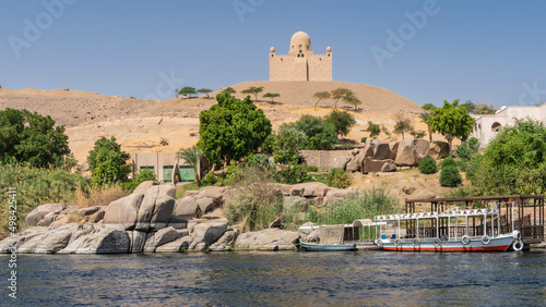 Tourist boats are moored near the river bank. Picturesque boulders at the water's edge. At the top of a sand dune, the old mausoleum of the Aga Khan is visible against the sky. Egypt. Aswan. Nile