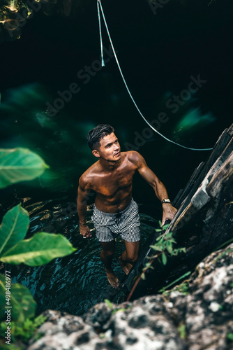 young tan male model climbing from a cenote in Tulum Mexico surrounded by green nature and a ladder