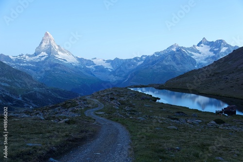 Morning scenery of majestic Matterhorn in blue twilight with a hiking trail winding thru rugged terrains & snow capped mountains reflected on peaceful Lake Stellisee in Valais, Zermatt, Switzerland